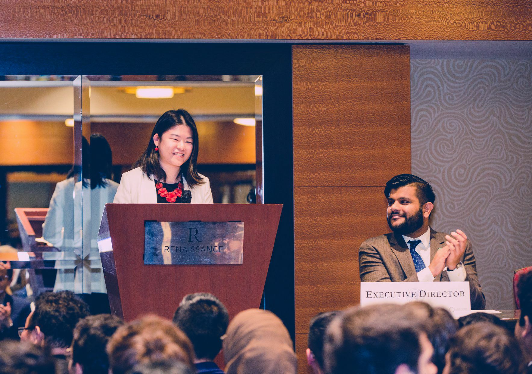 A speaker at the Renaissance Seattle Hotel podium with an applauding conference organizer